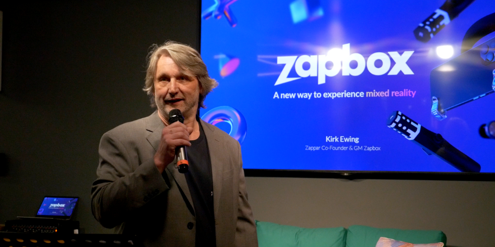 Zapbox General Manager Kirk standing on the stage with a microphone announcing our latest mixed reality announcements with a picture of the headset behind him.