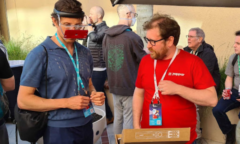 A photograph showing a man trying on Zapbox, the mixed reality headset with the box open in front of him. A Zappar team member stands next to him in a Zappar t-shirt talking him through.