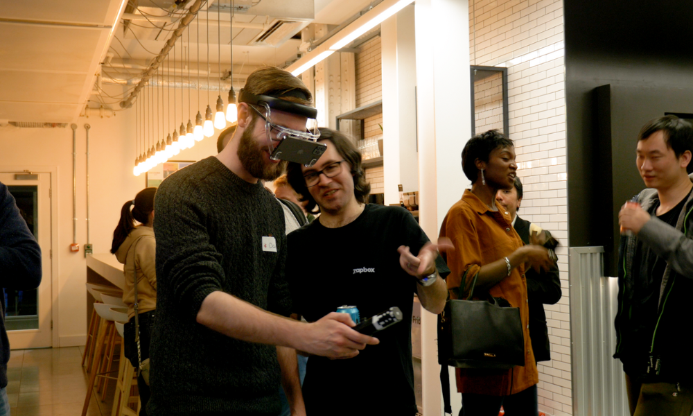 A man trys on zapbox, the mixed reality headset holding out the Bluetooth controller in front of him. Simon stands next to him gesturing with his hand. A group of people stand in the background drinking beer and chatting.